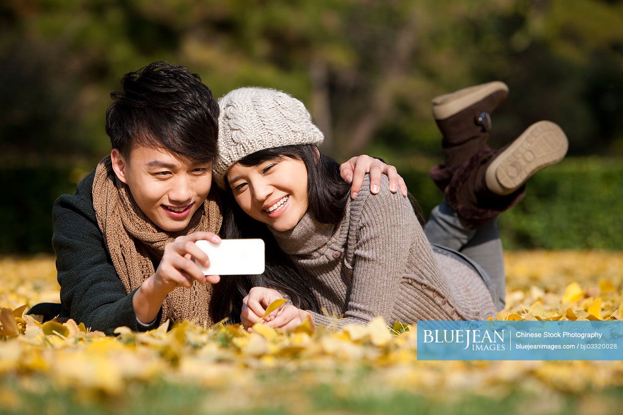Sweet young Chinese couple with smart phone lying on golden autumn leaves