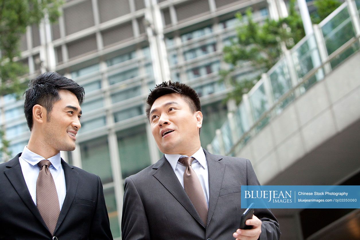 Happy Chinese coworkers walking and chatting with smart phone in hand outdoors, Hong Kong