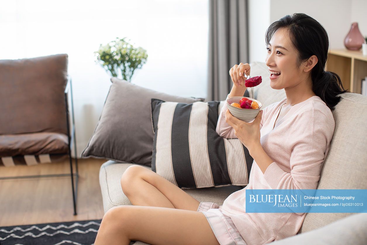 Happy young Chinese woman eating fruit salad in living room