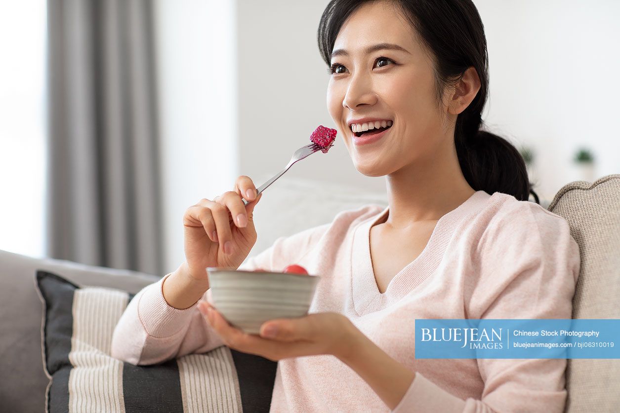 Happy young Chinese woman eating fruit salad in living room
