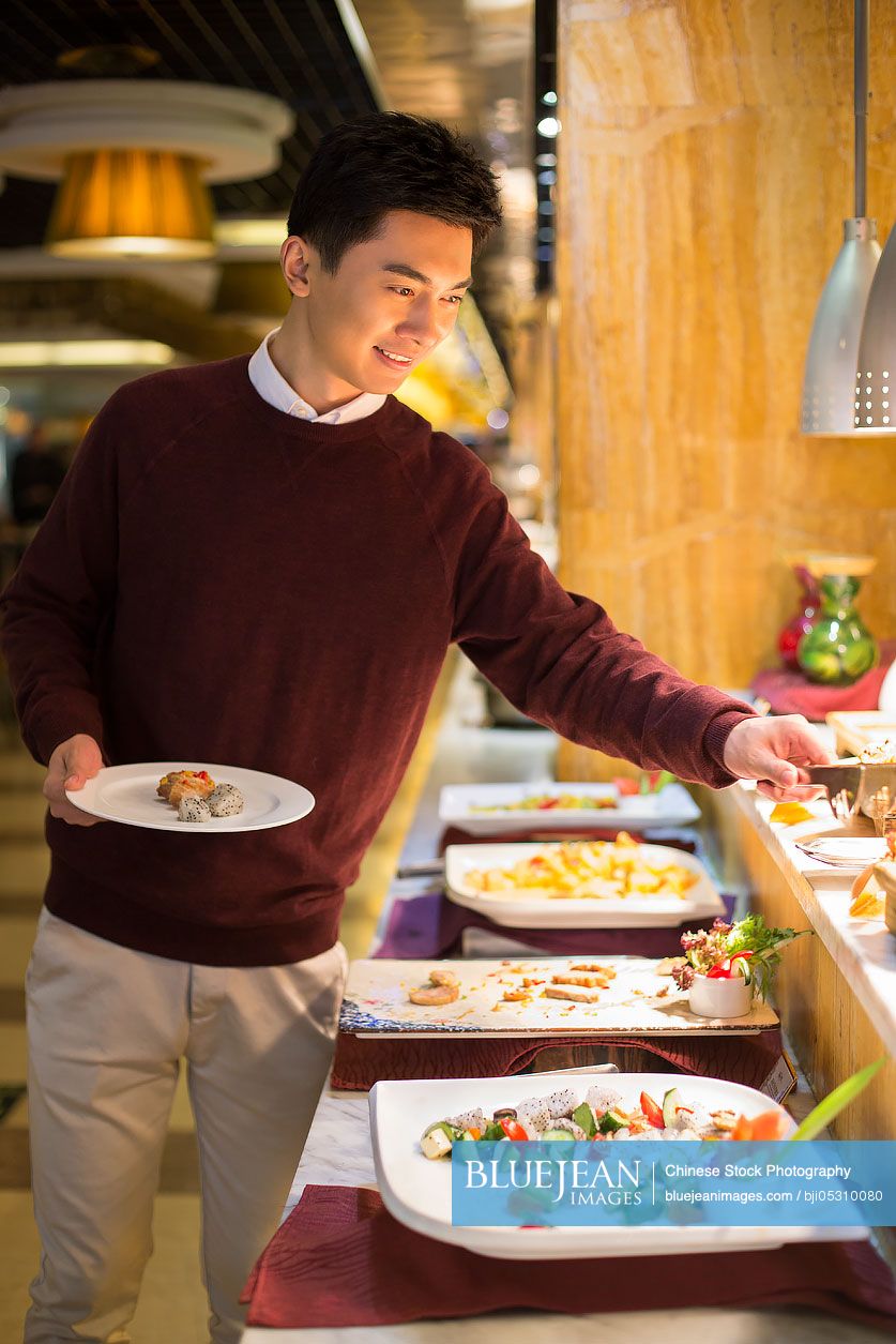 Cheerful young Chinese man taking food from buffet table