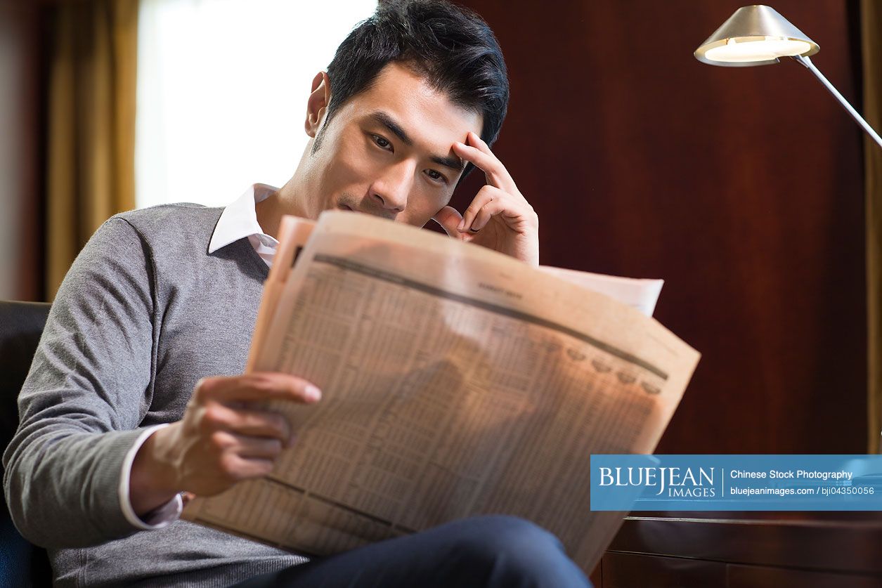 Young Chinese businessman reading newspaper in study