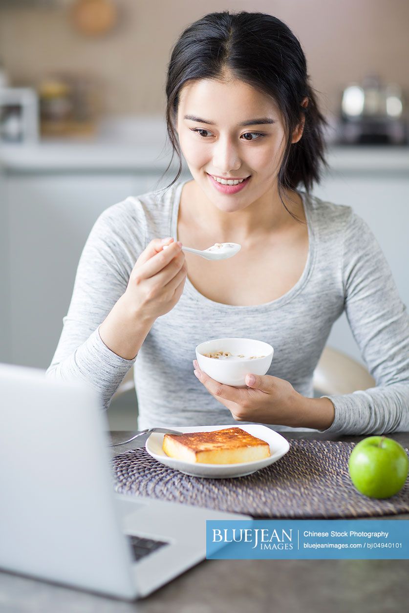 Young Chinese woman using laptop while having breakfast