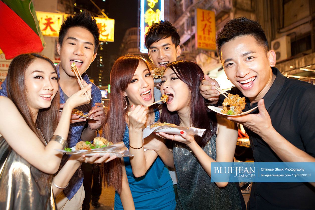 Cheerful young Chinese people trying some local snacks on Temple Street of Hong Kong, night view