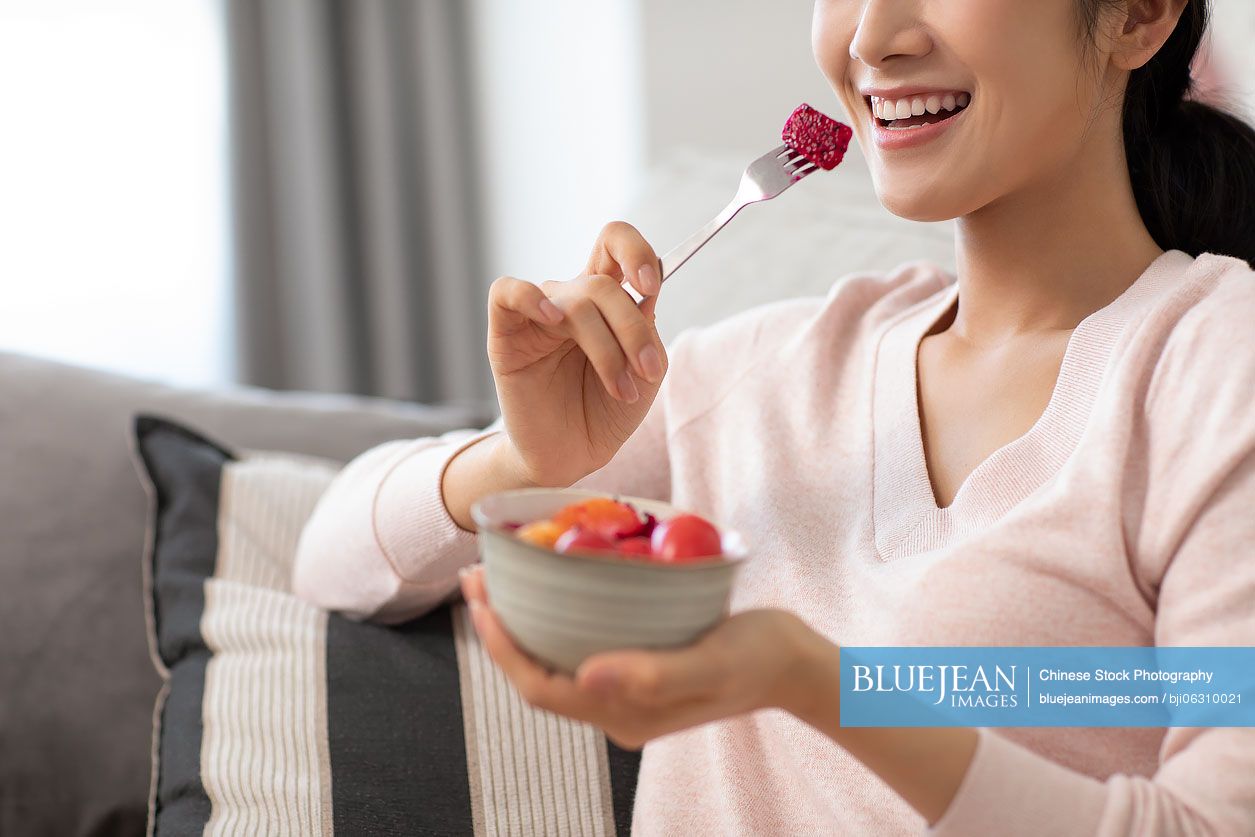 Happy young Chinese woman eating fruit salad in living room