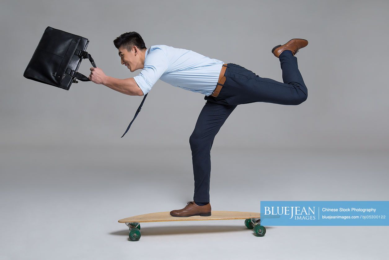 Cheerful young Chinese businessman skateboarding with briefcase