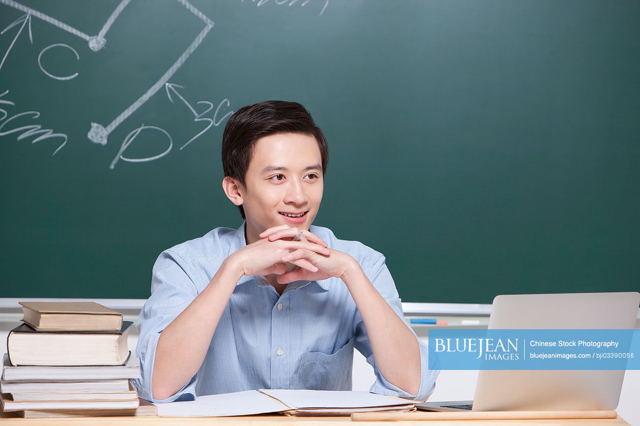 Cheerful male Chinese teacher hand on chin in classroom