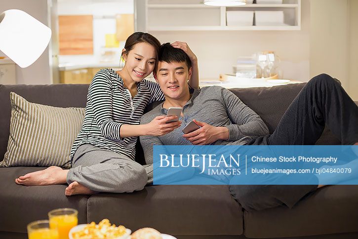 Young Chinese couple using smart phones on living room sofa