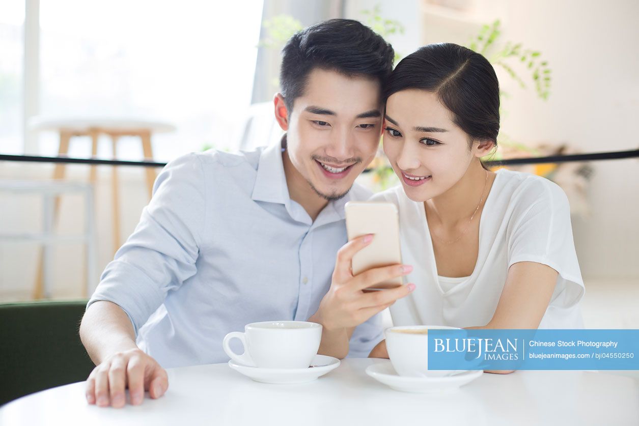 Happy young Chinese couple using smart phone in coffee shop