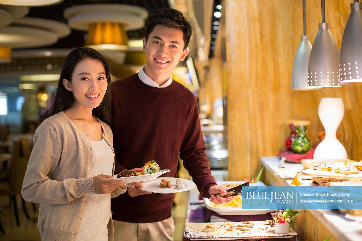 Cheerful young Chinese couple taking food from buffet table