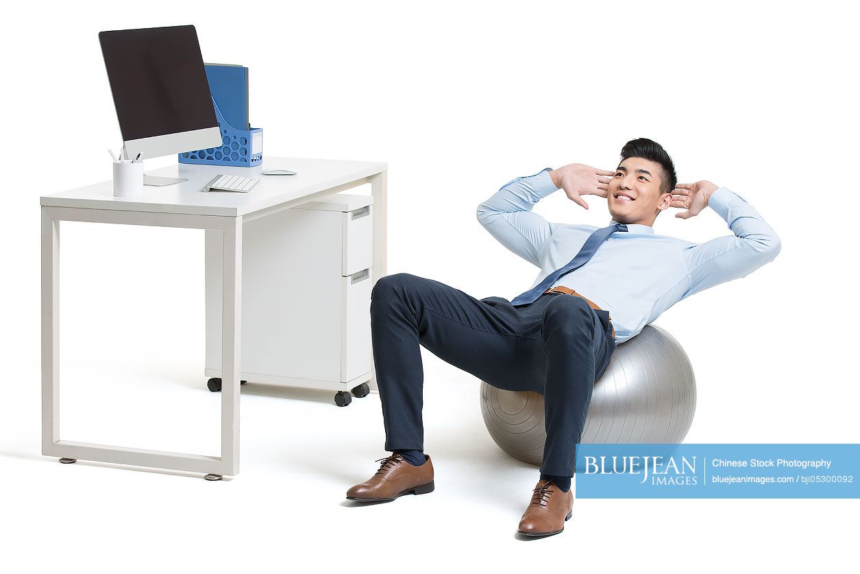 Young Chinese businessman exercising in office while sitting on fitness ball