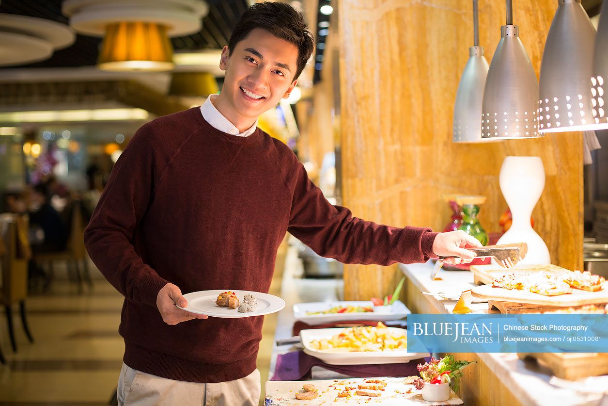 Cheerful young Chinese man taking food from buffet table