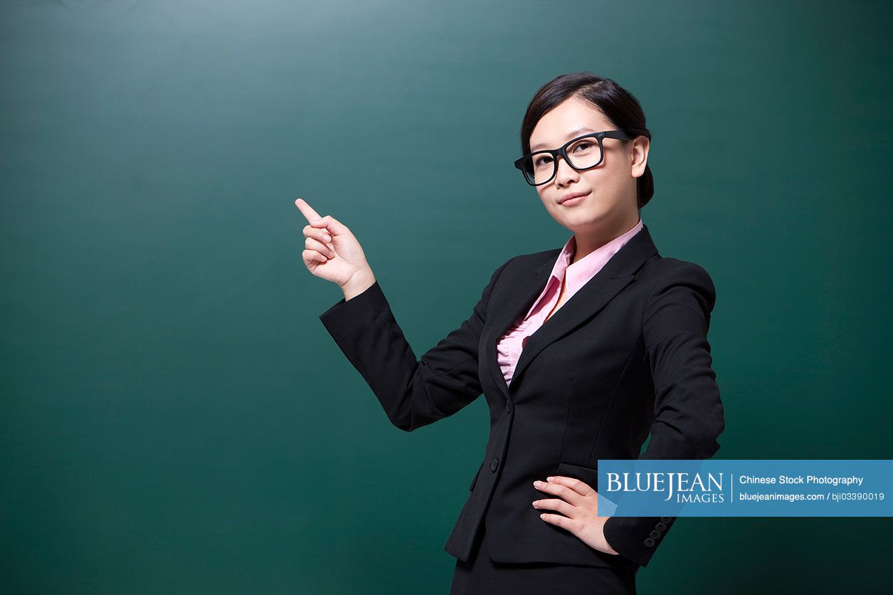 Serious female Chinese teacher pointing to blackboard in classroom