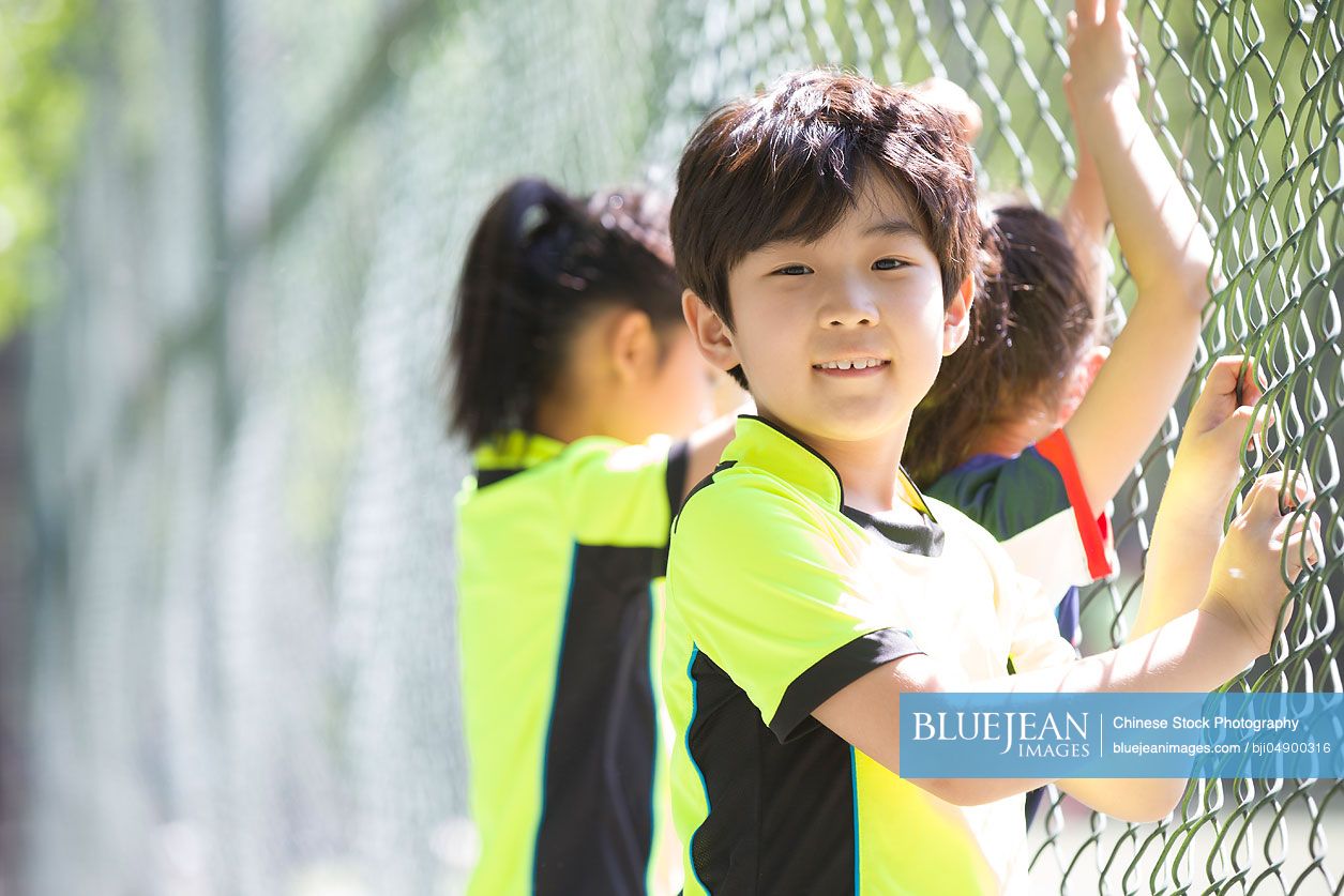 Happy Chinese children in sportswear leaning against chainlink fence