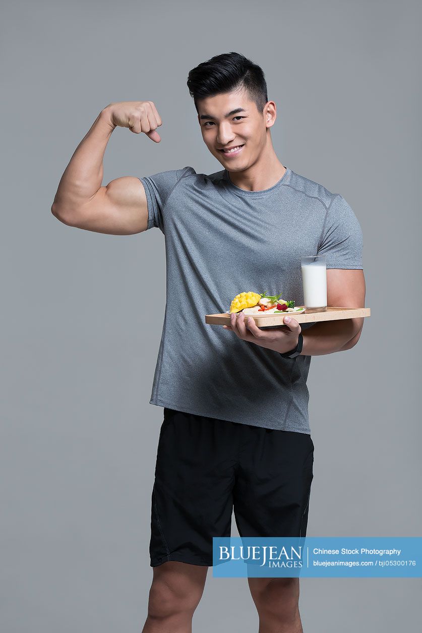 Young Chinese male athlete flexing muscles while holding vegetable salad and milk